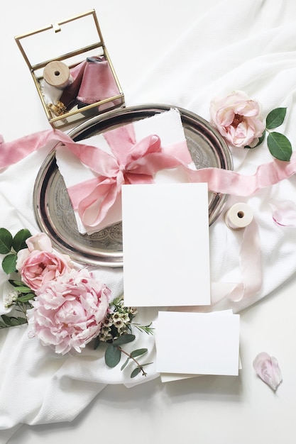 Vrouwelijke verticale bruiloft verjaardag mockup scène Blanco papier wenskaarten eucalyptus roze rozen pioen bloemen op witte tafel achtergrondGouden glazen doos met zijden linten Plat lag bovenaanzicht