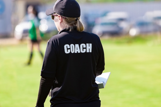 Vrouwelijke voetbaltrainer in zwart coach-shirt op een sportveld in openlucht, kijkend naar haar teamspel