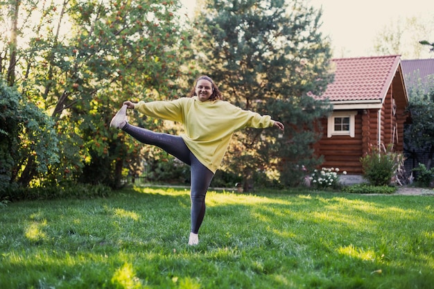 Vrouwelijke volwassene glimlachend op groen gras doen oefeningen in sportkleding met één voet omhoog op achtertuin vol zonlicht met landhuis en hoge bomen op de achtergrond Lichaam positief