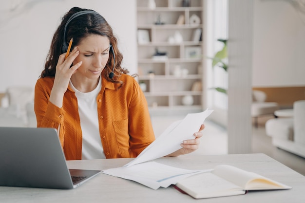 Vrouwelijke werknemer in headset kijkt door papieren document die thuis op laptop werkt Werken op afstand