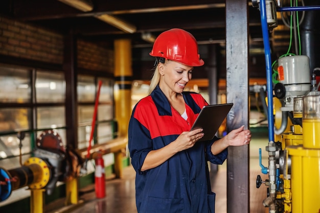 Vrouwelijke werknemer van middelbare leeftijd in werkkostuum met helm op hoofd die tablet gebruikt om op druk op machine te controleren terwijl hij in verwarmingsinstallatie staat.