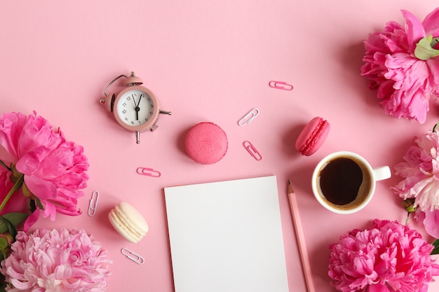 vrouwelijke werkplek met notitieblok potlood wekker pioenroos bloemen kopje koffie en macarons