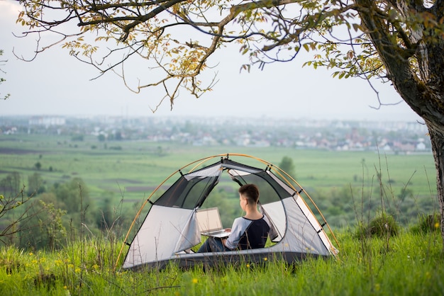 Vrouwelijke zitting in een tent bovenop een heuvel die haar laptop met behulp van terwijl het kamperen