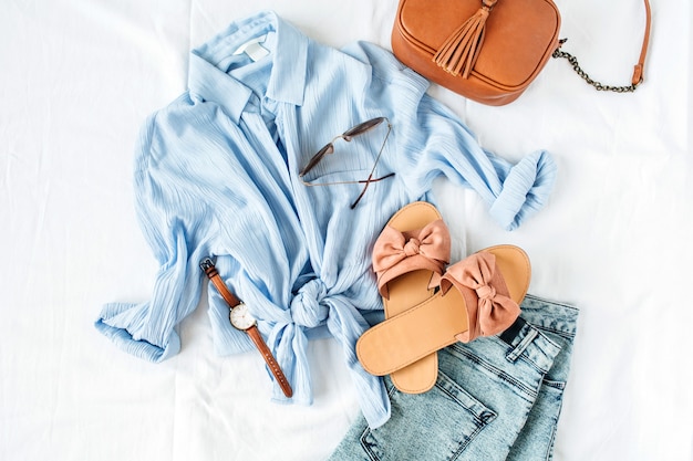 Vrouwelijke zomermode-compositie met blouse, pantoffels, tas, zonnebril, horloge en korte broek op wit oppervlak