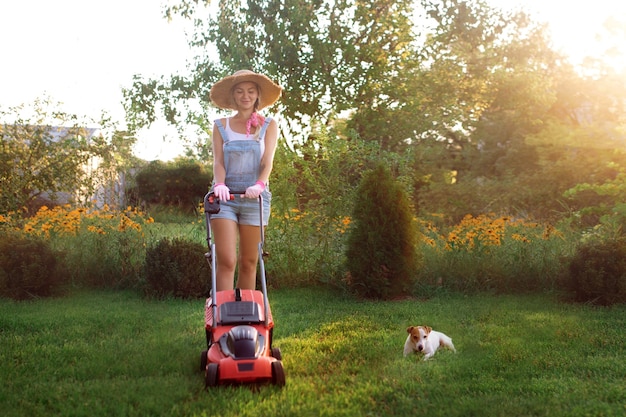Vrouwen tuinman. Een sexy meisje met een hoed maait het gras in de buurt van het huis met een grasmaaier. Landschapsontwerp en gazononderhoud