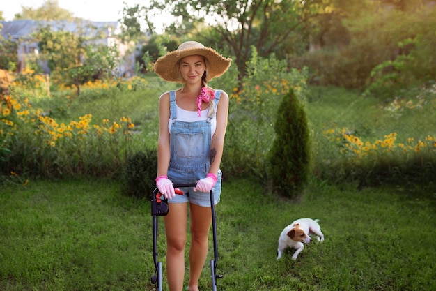 Vrouwen tuinman. Een sexy meisje met een hoed maait het gras in de buurt van het huis met een grasmaaier. Landschapsontwerp en gazononderhoud