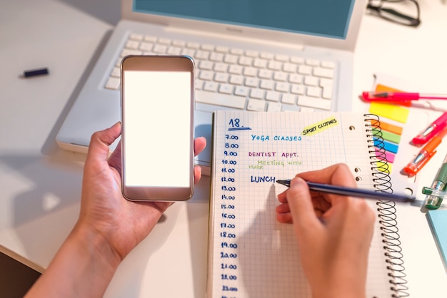 Vrouwenhand die in agenda schrijven die een mobiele telefoon op een bureau thuis raadplegen of bureau