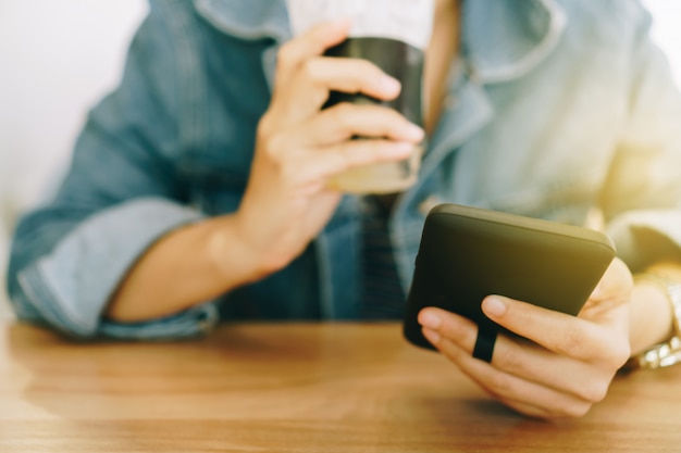 Vrouwenhand die smartphone gebruiken terwijl het drinken van koffie met de achtergrond van de koffiewinkel.