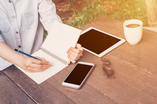 Vrouwenhand het schrijven notitieboekje en telefoon, tablet op lijst in tuin bij koffiewinkel met gestemde wijnoogst.