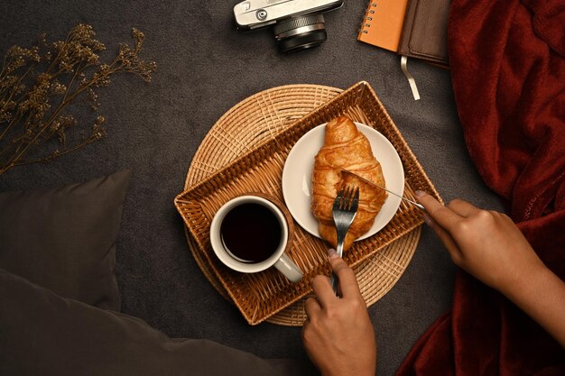 Vrouwenhanden die een stuk croissant met mes en vork snijden voor ontbijt met koffiekop op houten dienblad