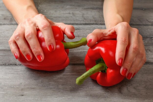 Vrouwenhanden die twee rode groene paprika's houden die op grijs houten bureau worden geplaatst.