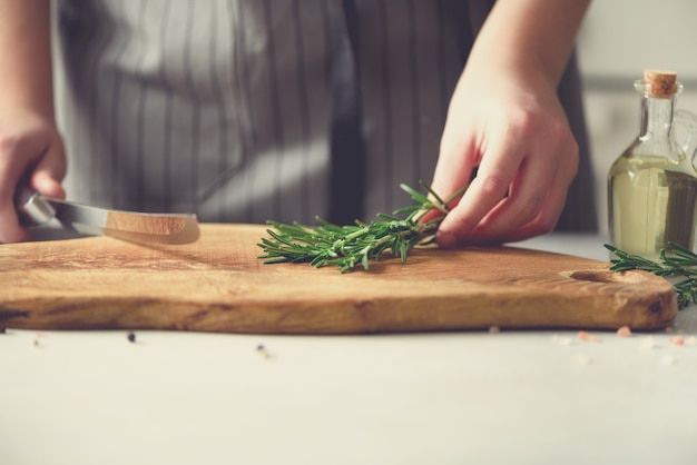 Vrouwenhanden die verse groene rozemarijn op houten hakbord in binnenlandse keuken snijden,. Ruimte kopiëren. Eigengemaakt voedsel conceplt, gezond recept. Breng me naar het werk