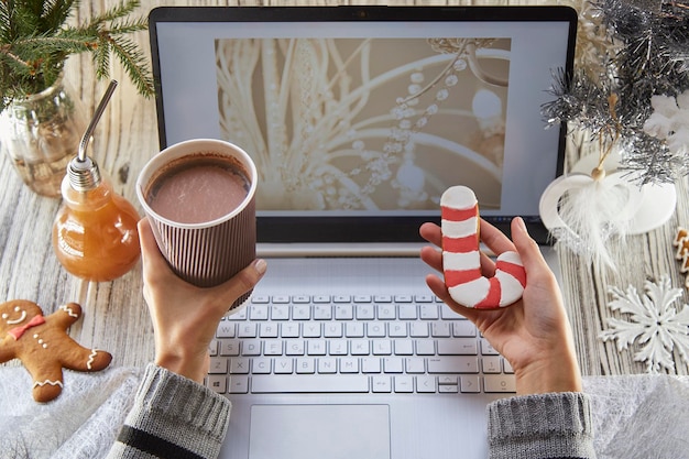 Vrouwenhanden houden een kartonnen kopje cacao en peperkoek vast Feestelijk thuiskantoor laptopmodel versierde kerstboom Werk op afstand kijken naar webinar, studeren of familievrienden bellen