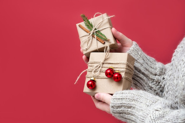 Vrouwenhanden houden geschenkdozen van kraftpapier vast met kerstdecor op een rode achtergrond, close-up. nieuwjaar achtergrond. geschenkverpakkingen van natuurlijke materialen in retro, vintage, trendstijl.