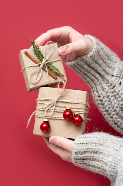 Vrouwenhanden houden geschenkdozen van kraftpapier vast met kerstdecor op een rode achtergrond, close-up. Nieuwjaar achtergrond. Geschenkverpakkingen van natuurlijke materialen in retro, vintage, trendstijl.