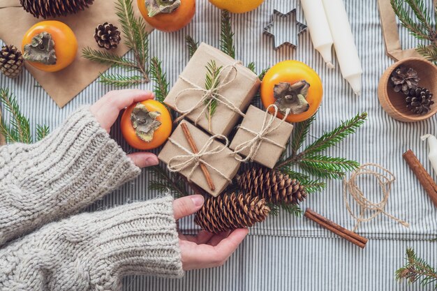 Vrouwenhanden houden geschenken vast voor het nieuwe jaar. Kerst achtergrond met geschenkdozen, kegels, kerstboom en persimmon, bovenaanzicht, close-up. Met de hand gemaakt cadeau.