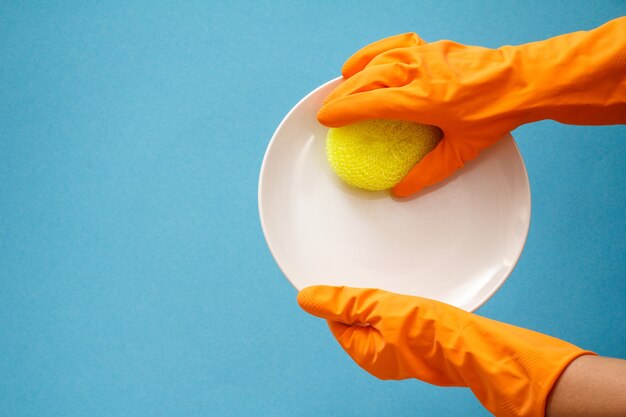 Vrouwenhanden in oranje rubberen handschoenen met witte plaat en gele spons op blauwe achtergrond. Was- en schoonmaakconcept.