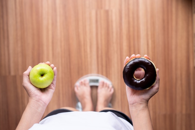 Vrouwenhanden met groene appel en gebakken chocoladedoughnut tijdens het staan op de weegschaal, gezonde voeding, dieetconcept