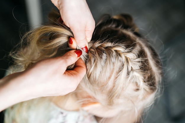 Vrouwenhanden weven vlechtjes voor een klein meisje, kinderkapsels thuis, een aartje haar.