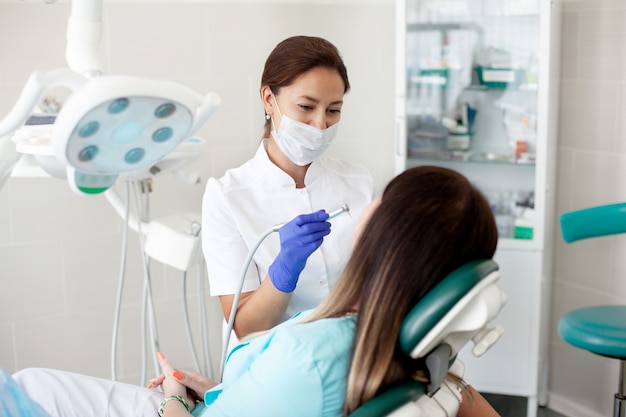 Vrouwentandarts behandelt een patiënt. Een vrouw in een masker zit in een tandartsstoel in de kliniek. Geneeskunde, gezondheid, tandheelkunde concept.