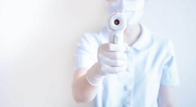 Vrouwenverpleegster die masker dragen en medische infrarode voorhoofdthermometer houden om lichaamstemperatuur voor virussymptomen te controleren.