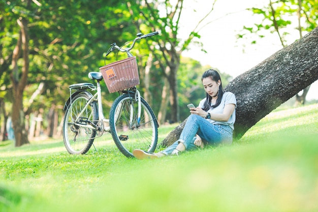 Vrouwenzitting in een park die op een boom leunen en aan muziek op hoofdtelefoons met een fiets luisteren