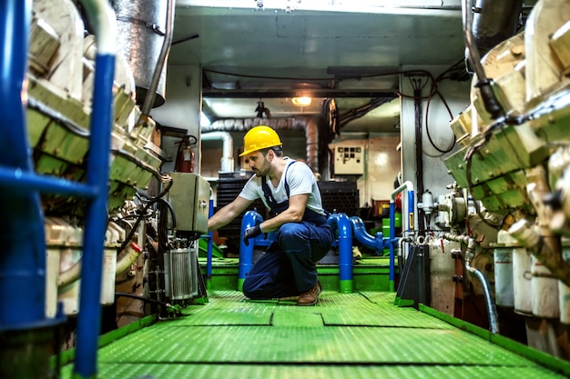 Foto waardig kaukasisch monteur in overall en met helm geknield in schip en motor repareren