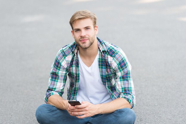 Wacht op oproep straatstijl man zit op de grond zorgeloze student vrije tijd besteden zomer mode knappe man geruit hemd mannelijke mode student ontspannen gebruik telefoon macho man chatten buiten