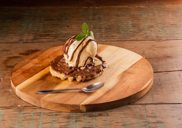 Wafel met chocolade en hazelnootroom en een bolletje vanille-ijs smeltend op een rond houten bord met dessertlepel hoekaanzicht