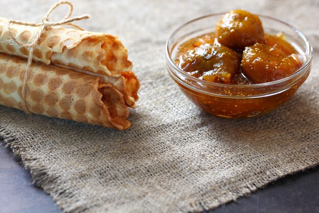 Waffle rolls three pieces tied with rope and fig jam in a glass bowl on burlap and on dark countertop. Gorizontal frame.