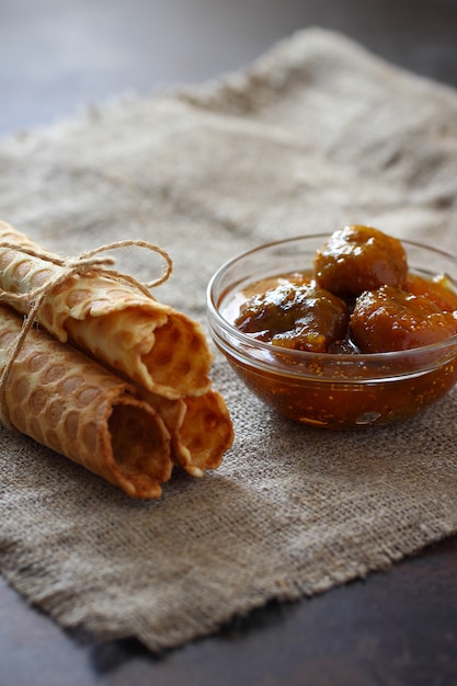 Waffle rolls tied with rope three pieces and fig jam in a glass bowl on burlap and on dark countertop.