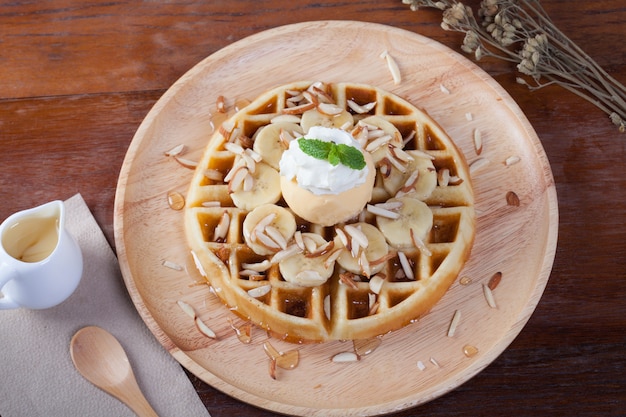 Waffle with banana and ice-cream vanilla on top has whip cream and peppermint sprinkle with almonds around.