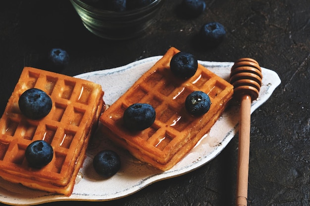 Waffles are laid out on plate, decorated with blueberries, doused with honey from honey spoon.