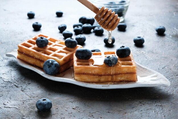 Waffles are laid out on plate, decorated with blueberries, doused with honey from honey spoon.