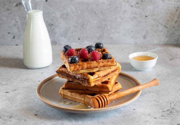 Waffles with fresh raspberries blueberries honey and bottle of milk on a concrete background Side view horizontal