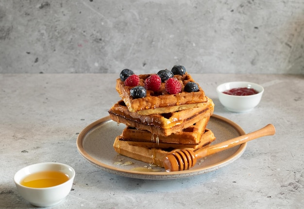 Waffles with fresh raspberries blueberries honey and wooden spoon on a concrete background Side view horizontal