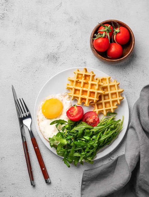 Waffles with fried egg cherry tomatoes and arugula on a white plate on a gray texture background with spices and vegetables