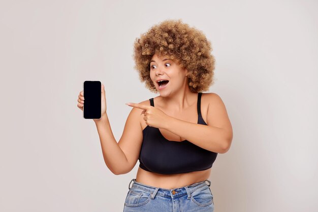 Photo waist shot of overjoyed excited young woman shows modern cellular with mockup display dressed in black lingerie bra and casual jeans isolated over white wall