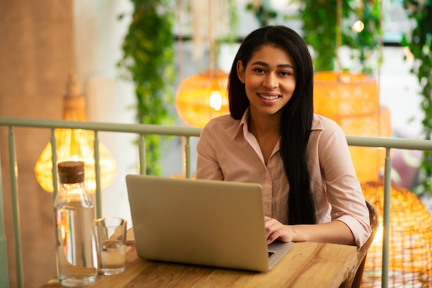 Mezzo busto di una giovane donna calma e contenta che sorride mentre lavora al suo laptop in un bar con una brocca di vetro d'acqua vicino al suo dispositivo