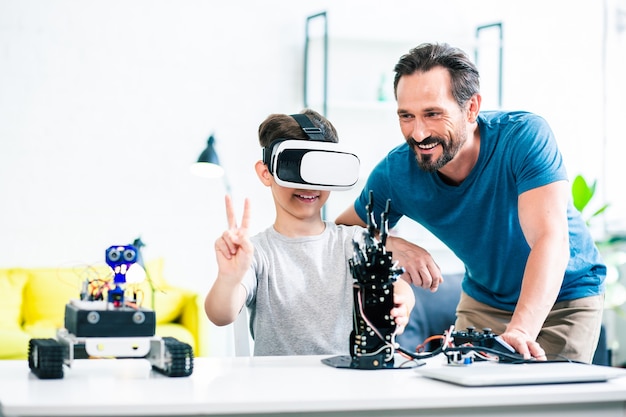 Waist up of a cheerful ingenious boy experimenting with his robotic device while his father standing nearby