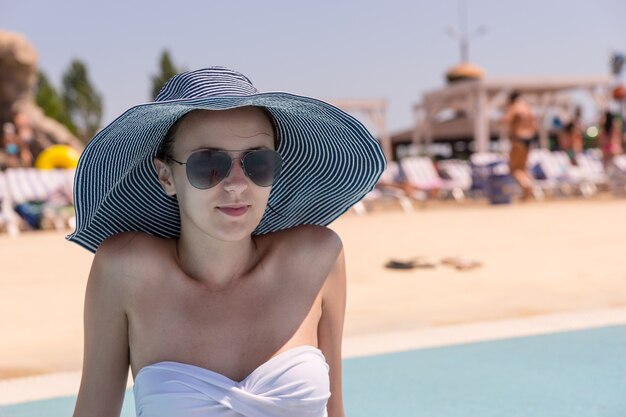 Waist Up Close Up of Young Woman on Vacation Wearing Sun Hat, Sunglasses and White Bikini While Sitting on Sunny Deck of Public Resort Swimming Pool