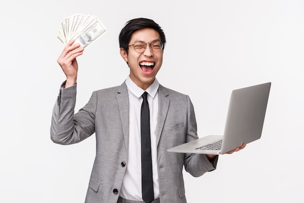 Waist-up of excited, lucky handsome rich asian guy, entrepreneur got his first money, selling or invest into company, shaking hand with dollars, big sum of cash, holding laptop and triumphing
