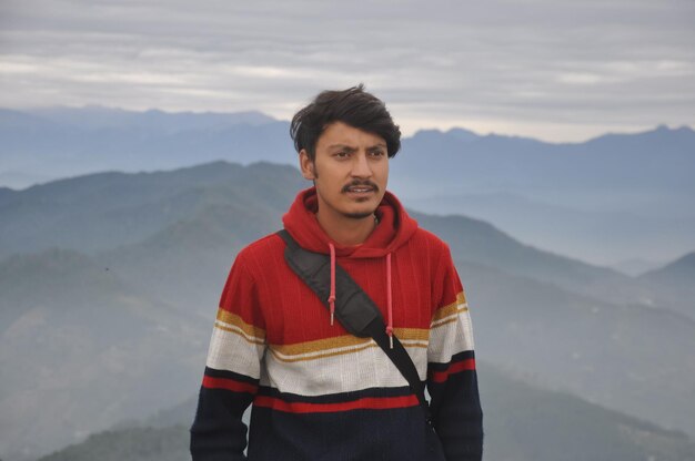 Photo waist-up of a indian young guy looking sideways while standing against the background of mountains