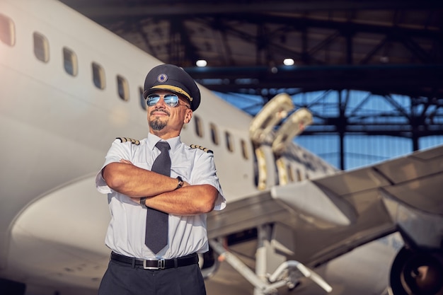 Photo waist up portrait of adult caucasian man keeping arms crossed while standing in front of airplane near the aviation hangar