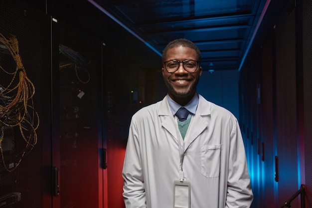 Waist up portrait of African American data scientist wearing lab coat and smiling at camera while working with supercomputer in server room, copy space