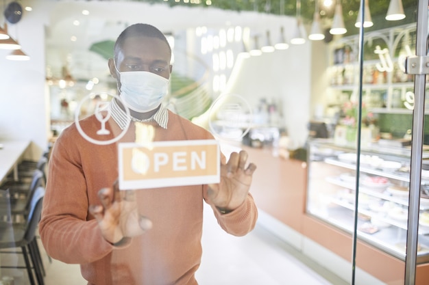 Waist up portrait of African-American man wearing mask while hanging OPEN sign on glass door of cafe in morning, copy space
