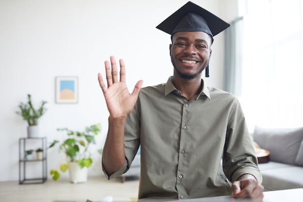 Waist up portrait of African-American young man wearing graduation cap and waving at camera in home interior, copy space