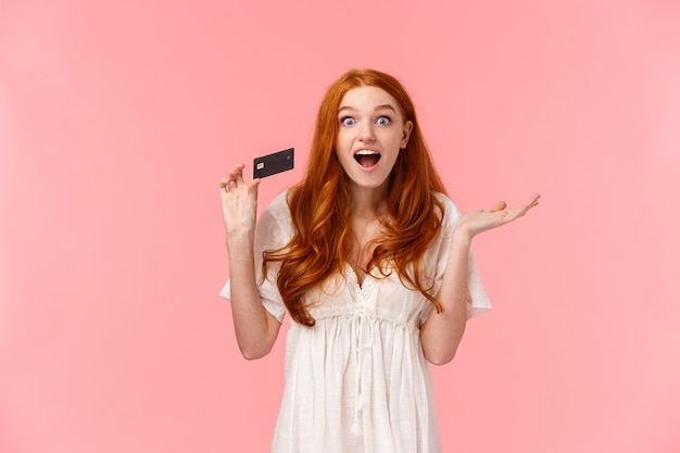 Waist-up portrait amused, wondered and excited fashionable feminine redhead girl telling about great promos and bonuses in bank, holding credit card and smiling astonished, pink wall