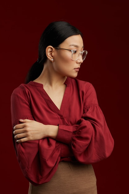 Waist up portrait of Asian woman wearing red blouse posing stiffly against maroon wall 