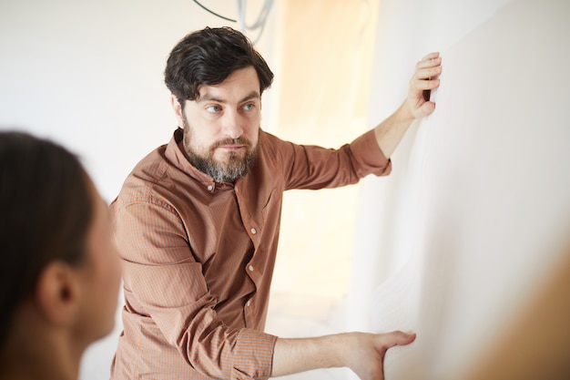 Waist up portrait of bearded man holding wallpaper to wall while redecorating house, copy space